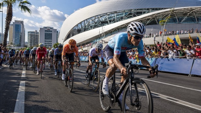 Carrera profesional de ciclismo en ruta en una moderna ciudad latinoamericana, con ciclistas compitiendo cerca de un estadio futurista y espectadores animando durante un evento deportivo diurno.